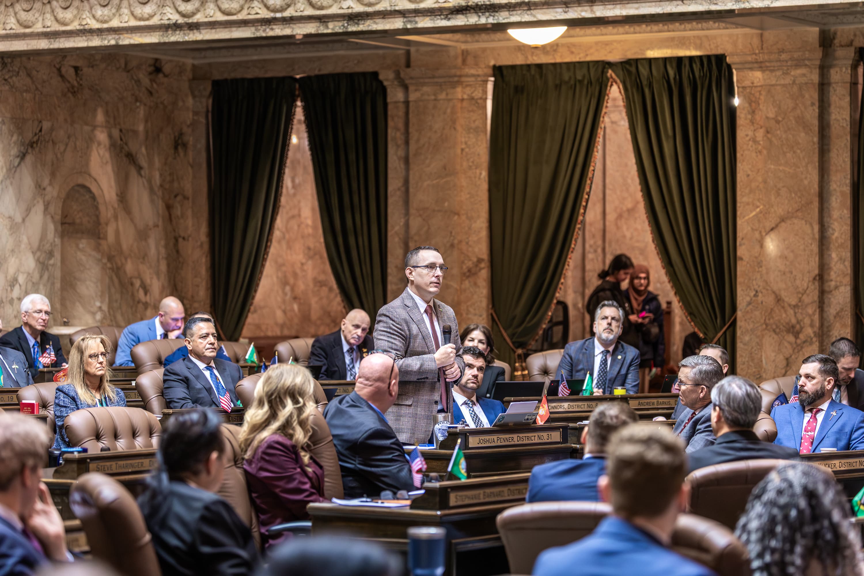 Representative Josh Penner speaking into a microphone on the House floor with colleagues nearby